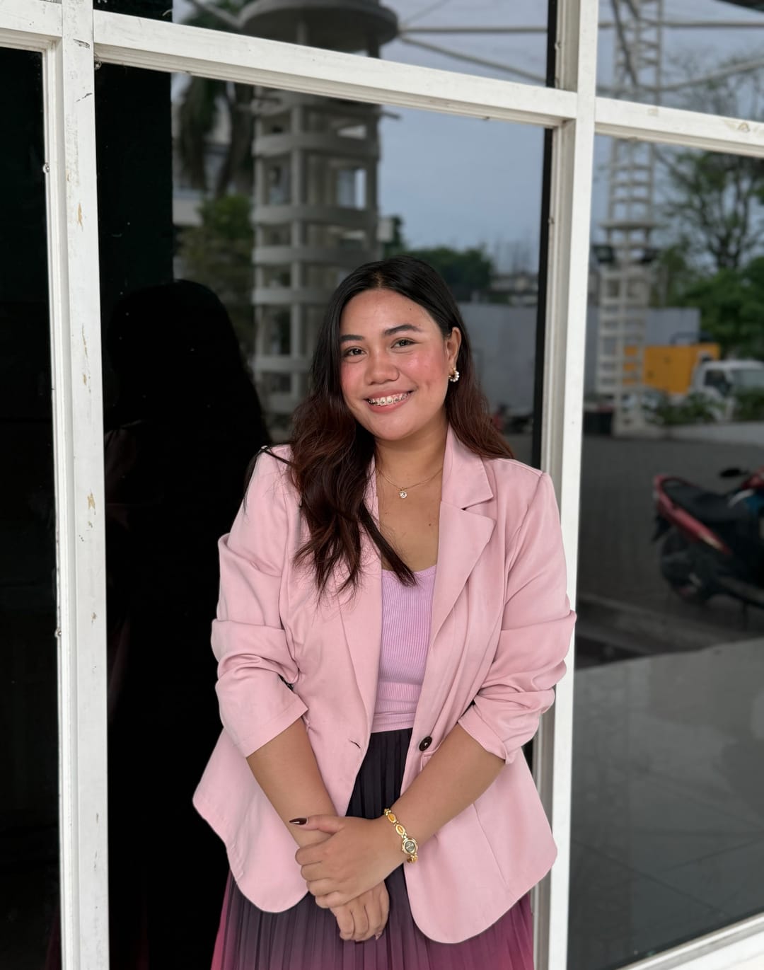Aliyha in a pink blazer and pleated skirt, standing in front of a white window frame